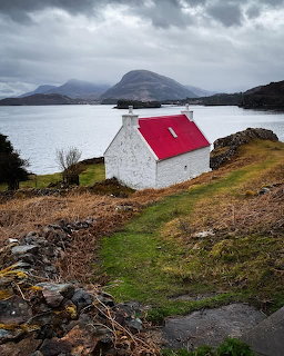 White cottage with red roof on Scottish loch shore with mountains and overcast sky