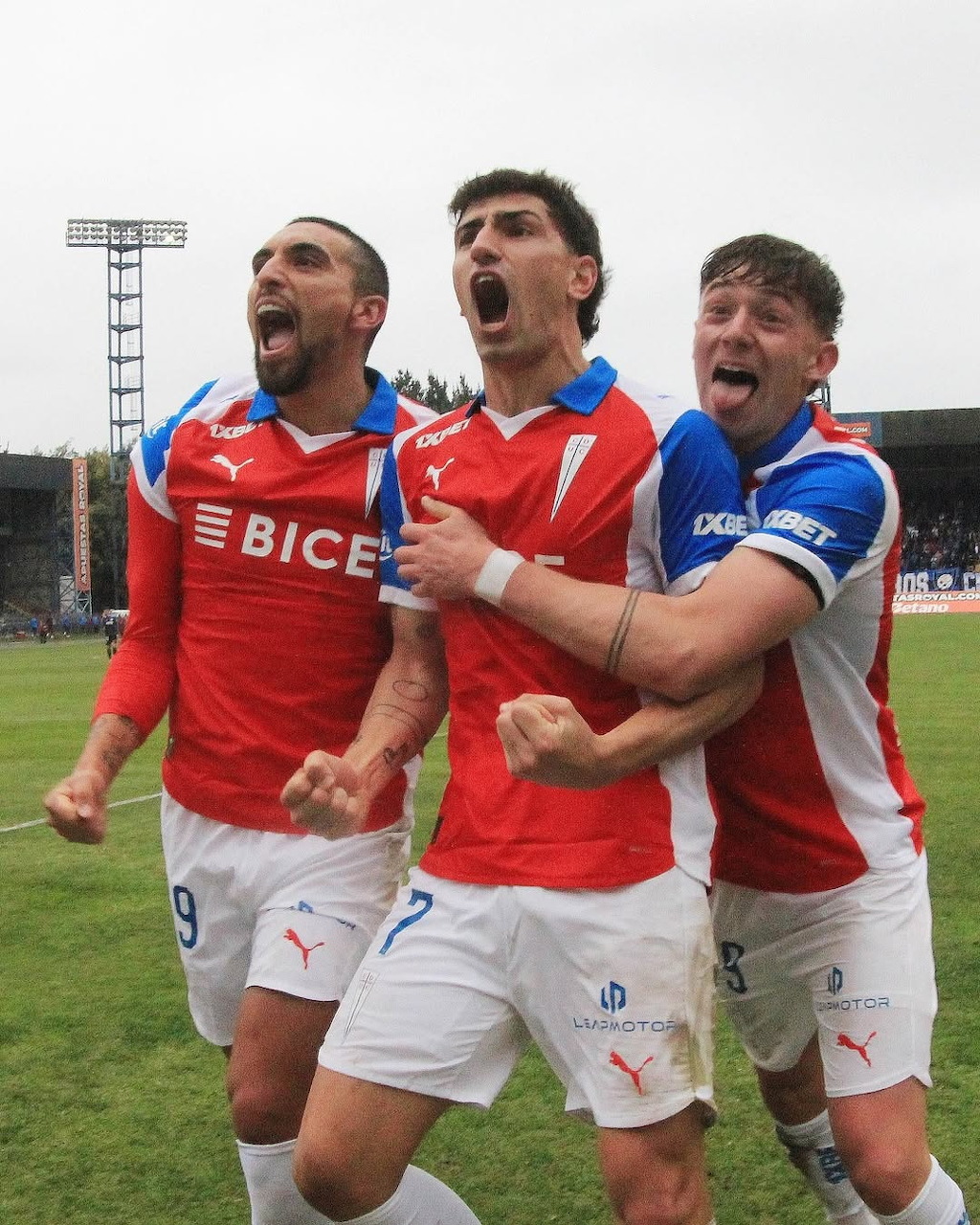 Three soccer players celebrating on a football field in red, blue, and white kits