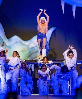 Female performer standing on platform above dancers in white outfits under blue stage lighting