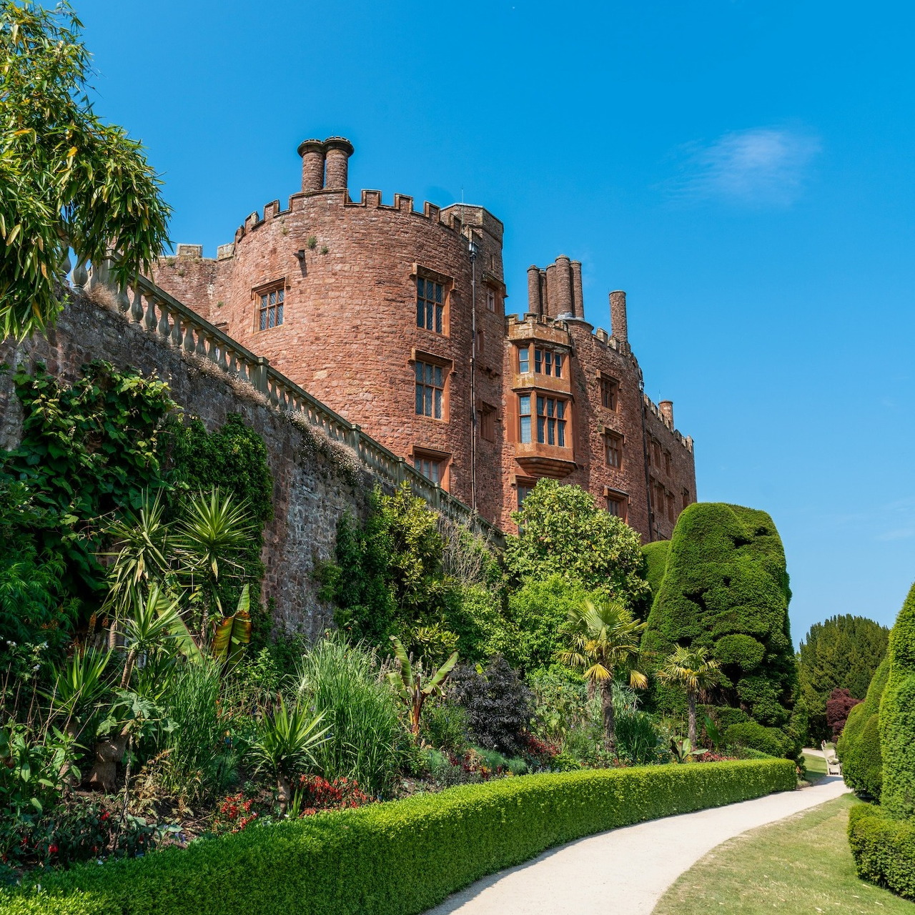 Red brick castle with formal terraced gardens, sculpted hedges, and curved path on a sunny day