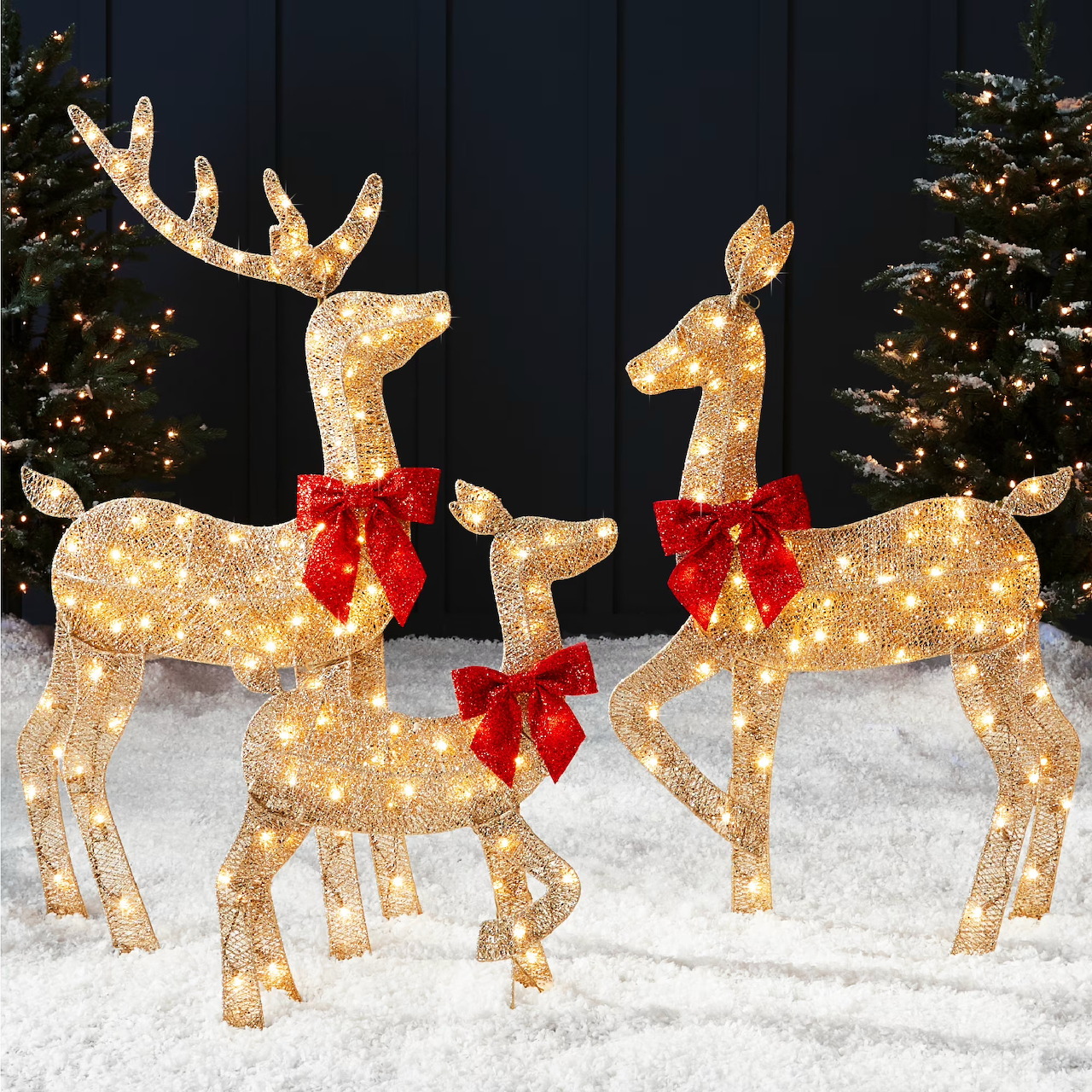 Three illuminated gold reindeer lawn ornaments with red bows on snowy ground beside Christmas trees