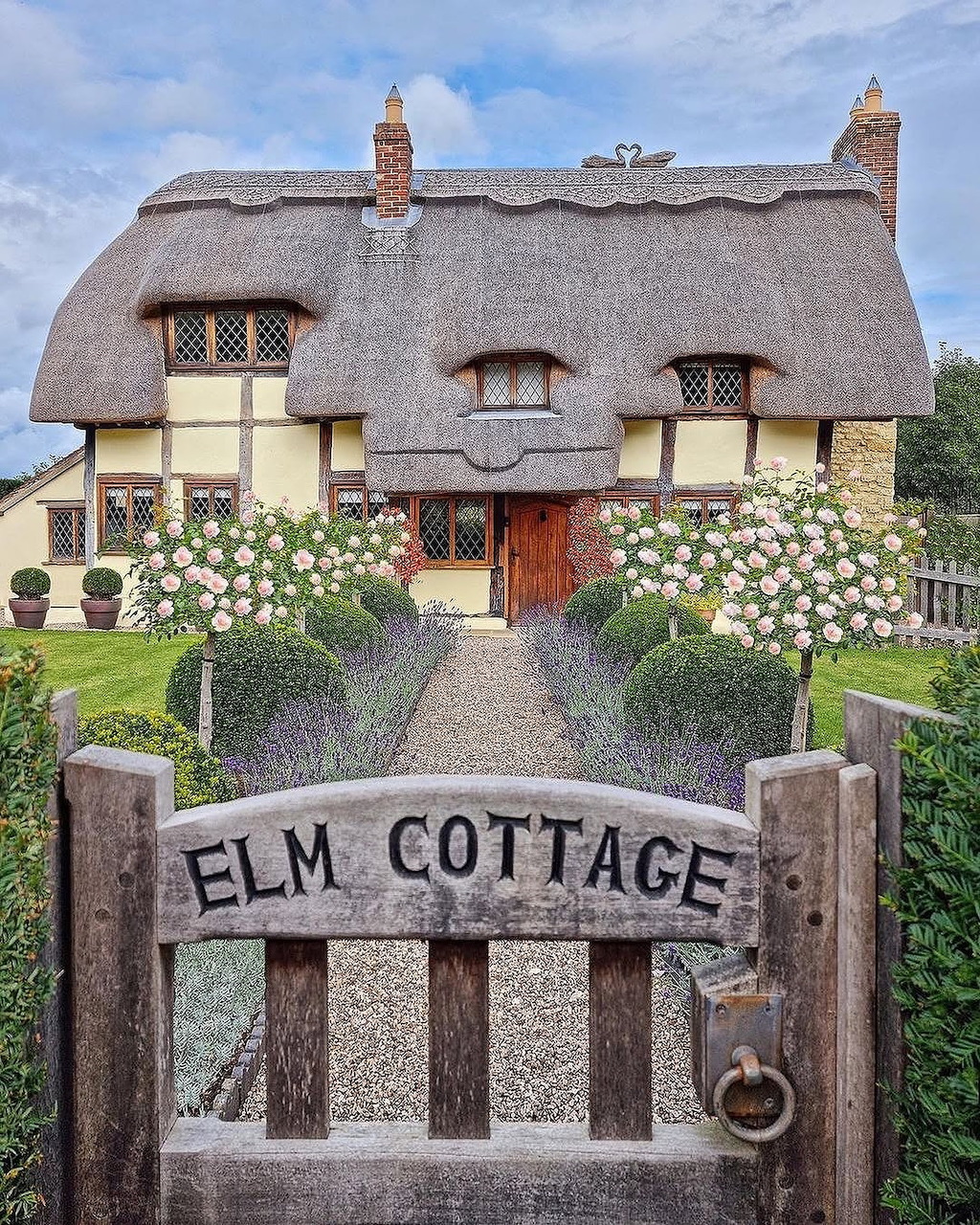Thatched cottage with wooden gate reading Elm Cottage, surrounded by flowering garden and blue sky