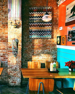 Bright taco restaurant interior with exposed brick, zigzag tapestry, turquoise counter and wooden furniture
