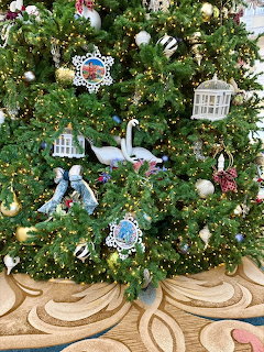 Decorated Christmas tree with white ornaments, blue bows, and miniature house decorations on ornate floor