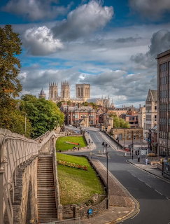 City street with stone steps, green lawns, and a cathedral in the distance under cloudy skies