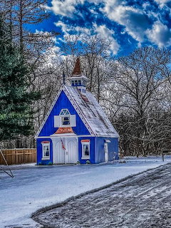 Small blue church in snow with bare trees and a bright cloudy sky