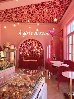 Pink cafe interior with floral ceiling, cupcake display case, velvet seating, and fairy lights