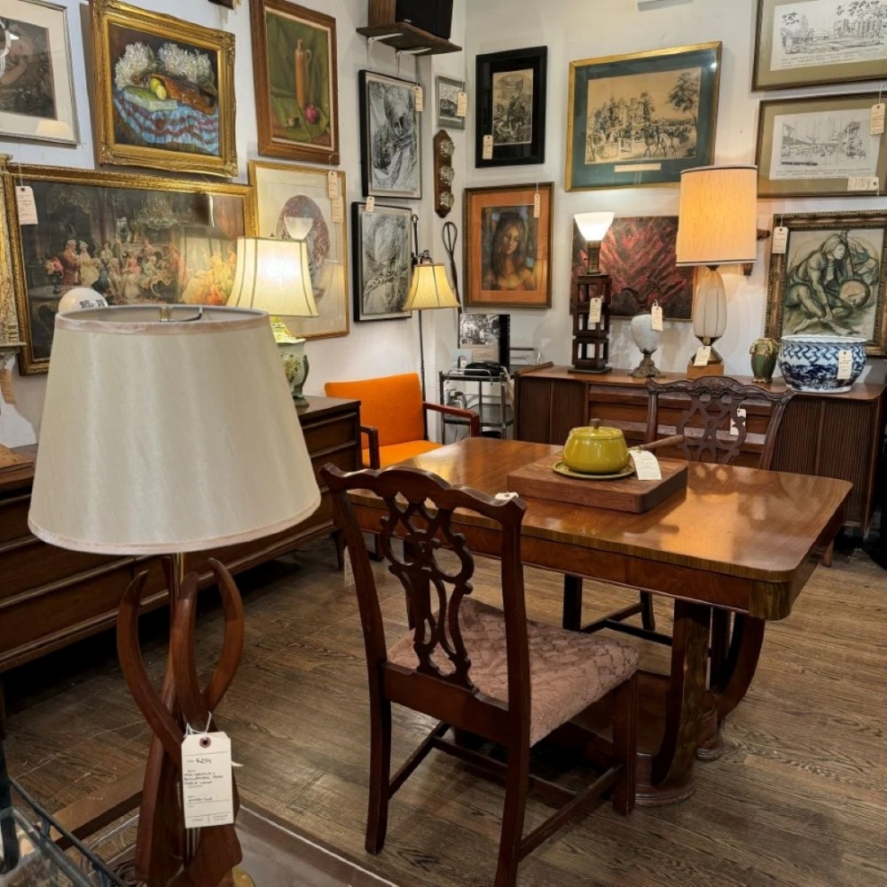 Antique store interior with wooden dining table, chairs, lamps, and framed artwork on walls