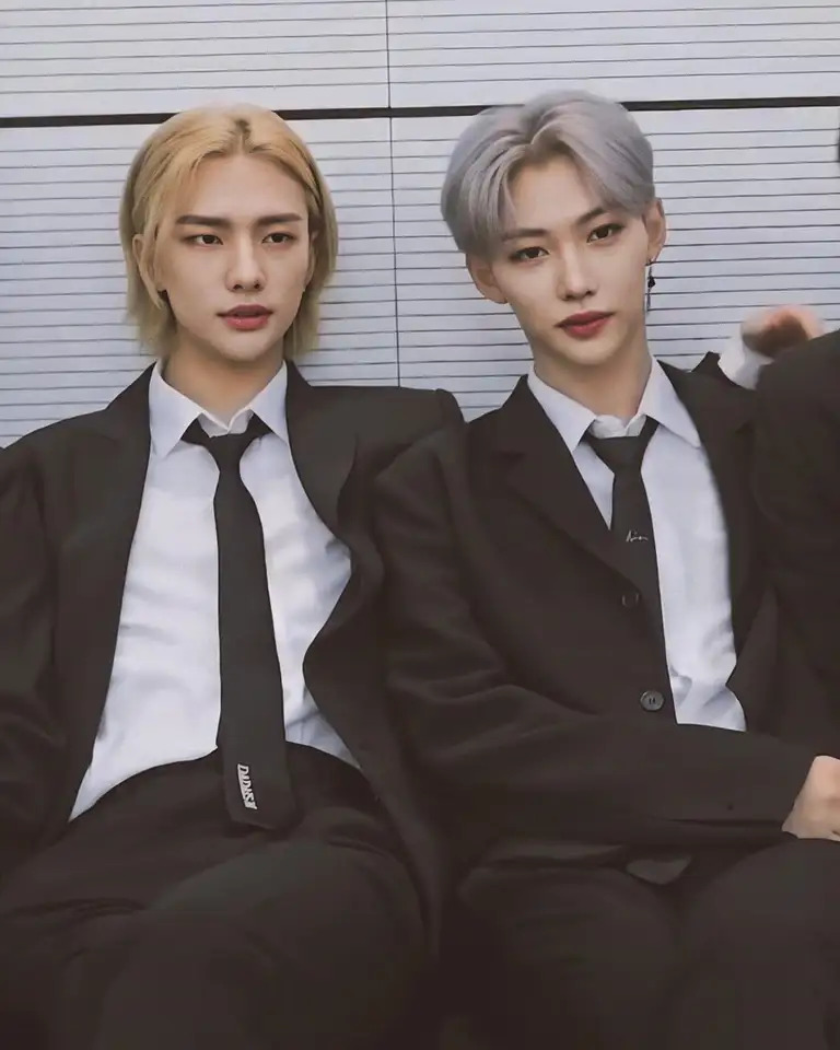 Two men in black suits and ties sitting together, one blonde and one with silver hair
