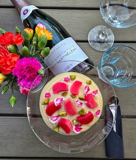 Yellow cake with rose petals, champagne bottle, glasses, and vibrant flowers on wooden surface