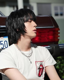 Young man with black hair and Rolling Stones t-shirt sitting near a dark car outdoors