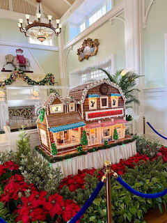 Large gingerbread house display indoors, surrounded by red poinsettias and holiday decorations