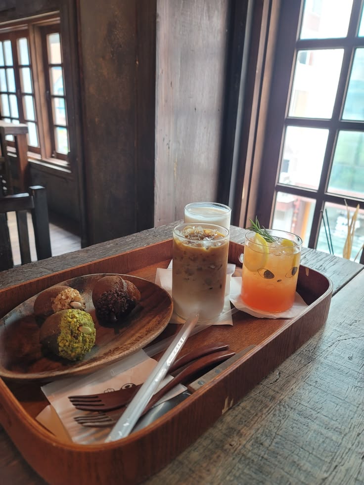 Wooden tray with iced coffee, orange drink, and pastries on a café table by a window