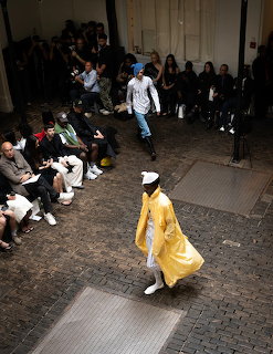 Overhead view of a model in yellow walking a coblestone runway with audience seated along the sides