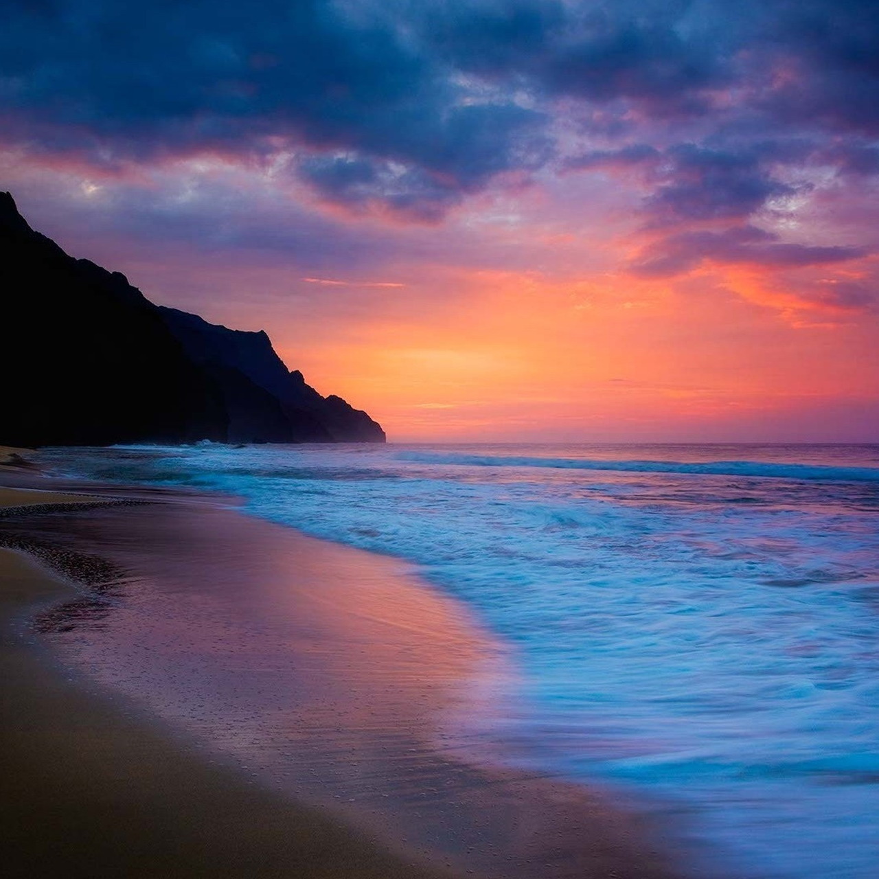Sunset over beach with rocky cliff, colorful sky and calm ocean waves reflecting on wet sand