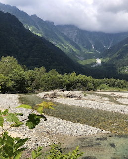 Mountain valley with river flowing through rocky bed, green forested slopes and cloudy peaks in background