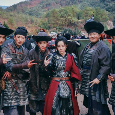 Group of actors in Chinese historical armor and red costume posing outdoors with green hills behind.