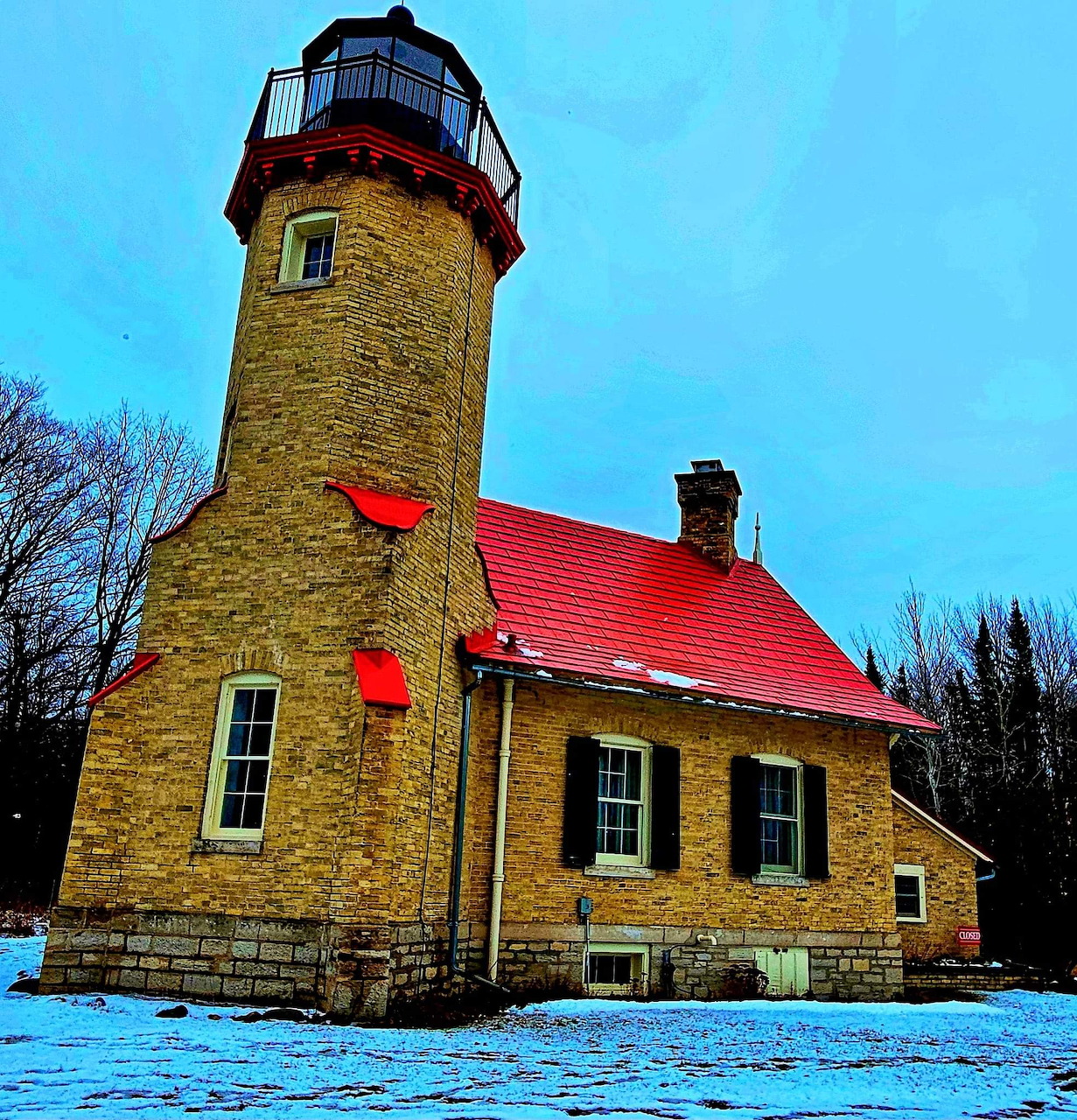 Yellow brick lighthouse with red roof in snow under blue sky
