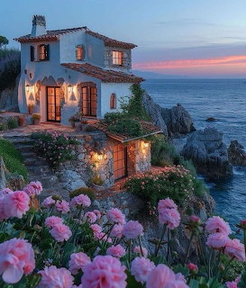 White Mediterranean cottage on rocky seaside cliff at sunset with pink roses in foreground