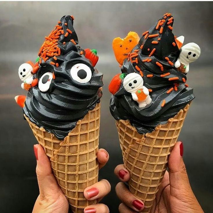 Two hands holding black soft-serve waffle cones decorated with candy eyes, sprinkles, and Halloween toppers