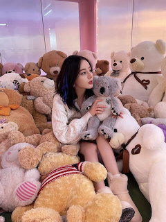 Young woman sitting among many stuffed teddy bears hugging a grey koala plush toy