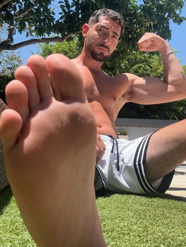 Muscular man sitting on grass flexing his arm with bare foot extended toward camera