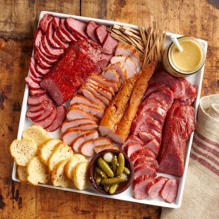 Tray of assorted sliced meats with bread, pickles, breadsticks, and mustard on a wooden table