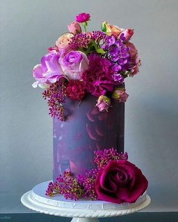Purple cake adorned with vibrant flowers and berries on white stand