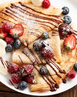 Stack of crepes with fresh berries, banana slices, powdered sugar and chocolate sauce on white plate