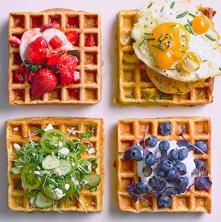 Four waffles with red strawberries, yellow fried eggs, green cucumber salad, and blue blueberries arranged in a square