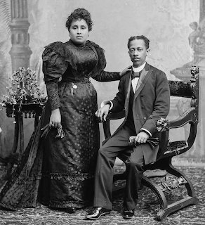 Black and white studio portrait of a couple in formal 19th century attire, woman standing and man seated