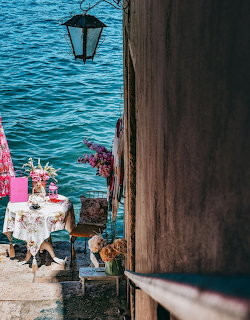 Small flower-covered table on a stone terrace beside blue sea with a hanging lantern and rustic wall.