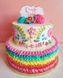 Two-tier cake with rainbow ruffles on bottom tier and colorful fondant flowers on top tier