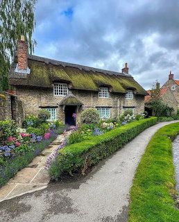 Stone cottage with green thatched roof and vibrant flower garden along country path