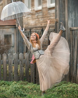 Woman in a flowing beige dress posing with a clear umbrella outdoors by a wooden fence