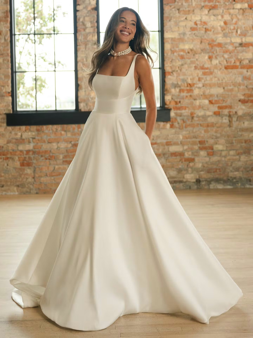 Smiling bride in white A-line satin wedding gown posing in front of brick wall with windows