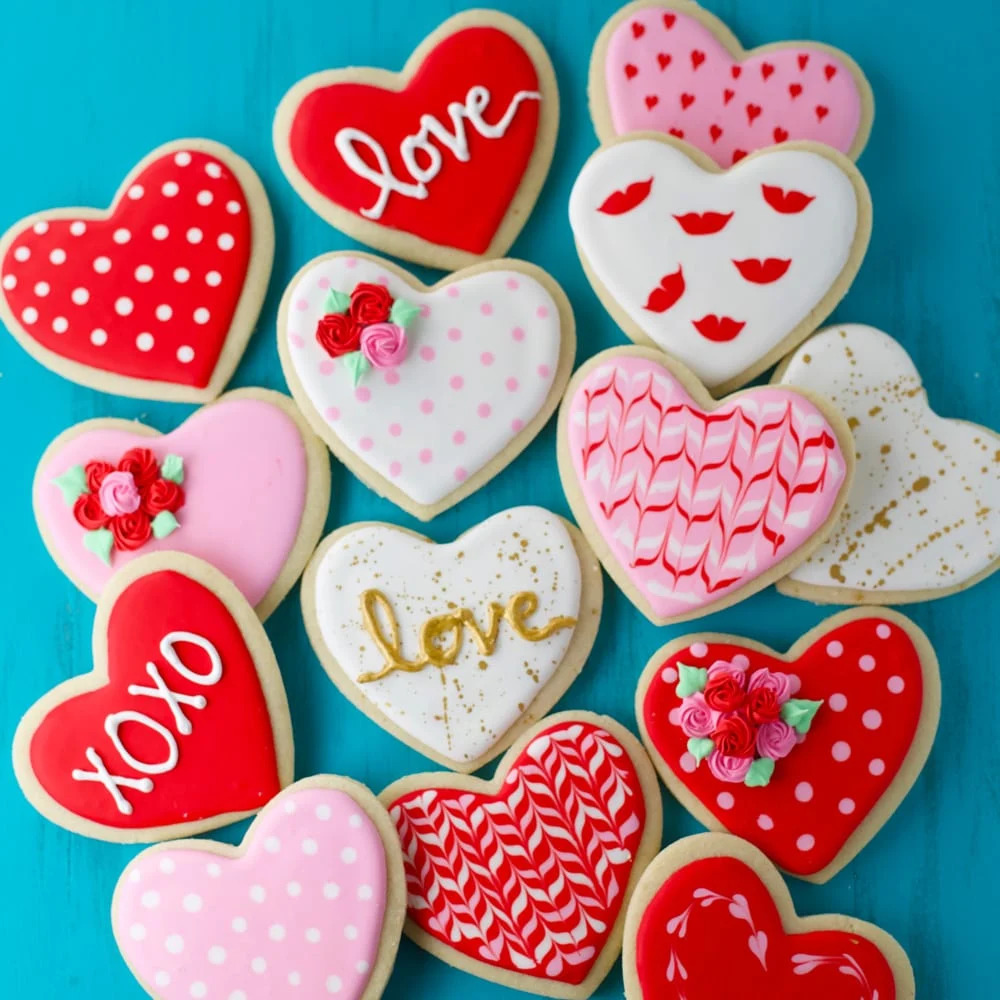 Assorted heart-shaped Valentine cookies with colorful icing designs including Love and XOXO messages on blue background