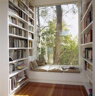 White built-in bookshelves surrounding a window seat reading nook with cushions and a view of trees