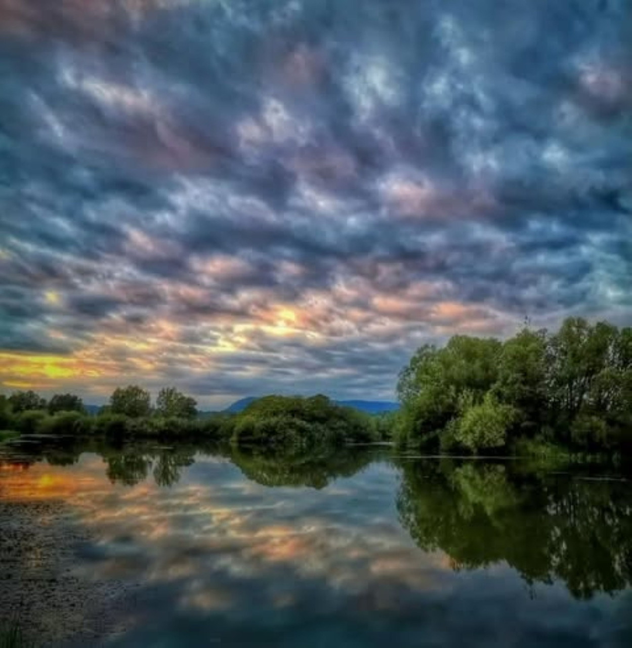Dramatic sunset clouds reflected in still lake water with trees along shore