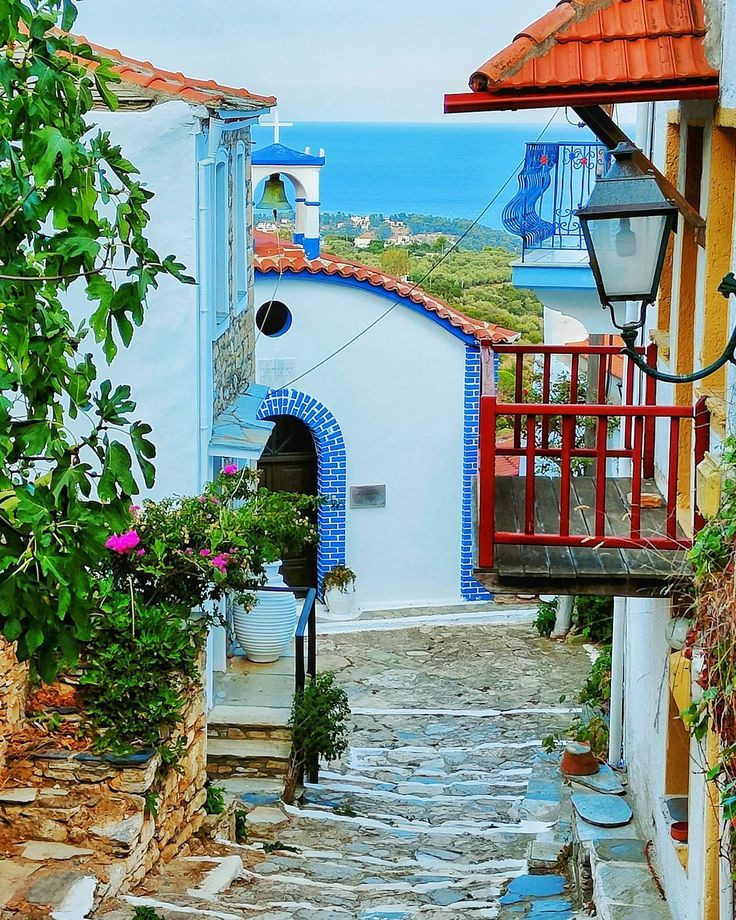 Narrow cobblestone street with white buildings, blue door, terracotta roofs, and sea view in Greek coastal village