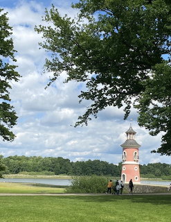 Pink lighthouse on green grass beside a lake with trees and cloudy sky