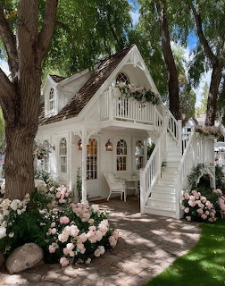 White cottage with arched windows and roses in a lush garden with trees and a stone path