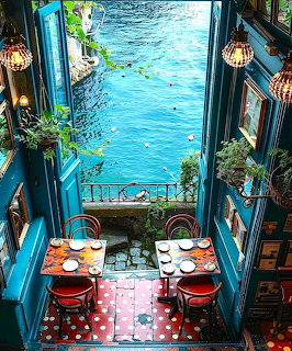 Overhead view of a colorful waterfront bistro with open doors, set tables, and turquoise sea beyond