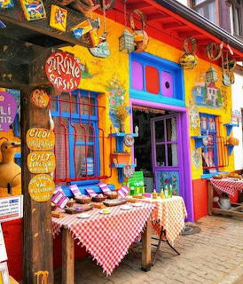 Brightly painted yellow cafe with blue windows, red canopy, hanging ornaments, and outdoor table with checkered cloth