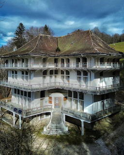 Old white mansion with balconies and a steep roof surrounded by trees under a cloudy sky