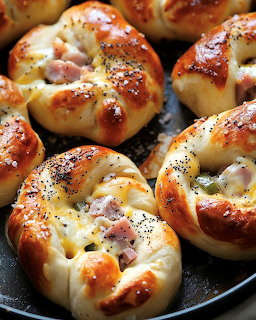 Golden baked pastry rolls stuffed with ham and cheese on a dark pan, topped with poppy seeds
