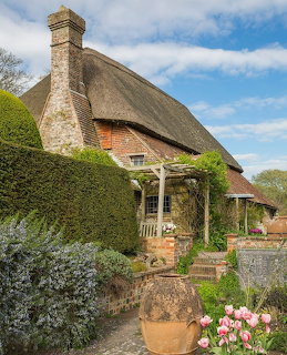 Thatched roof cottage with timber frame, stone chimney, and pink tulips in front garden under blue sky
