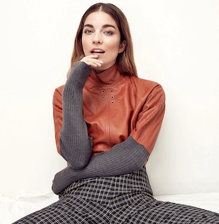 Woman in rust turtleneck and gray sleeves sitting against white background
