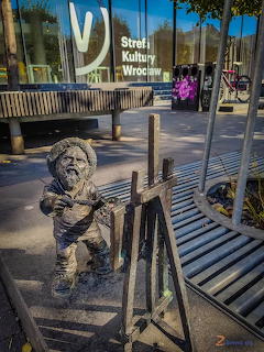 Bronze dwarf statue holding a palette and painting at a small easel on a metal bench in Wrocław