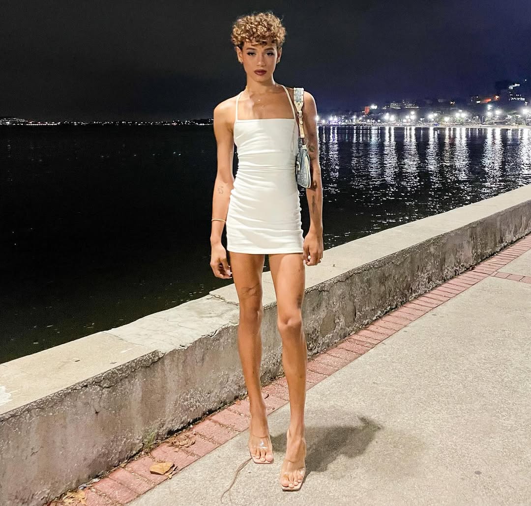 Woman in white dress standing on waterfront promenade at night with city lights reflecting on water